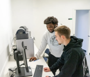 Two men work together at a computer station in a lab, both looking at the screen and interacting with the keyboard and mouse.